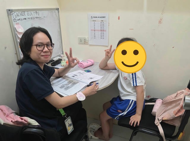 Seorang anak duduk di samping gurunya di ruang kelas 1:1, keduanya berpose tanda damai