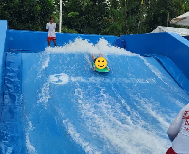 Seorang anak bermain papan di seluncuran ombak buatan di taman air