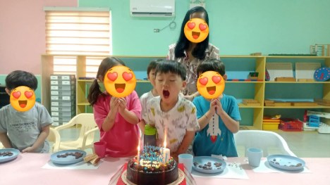 キンダーの教室でケーキのろうそくを吹き消す子どもと友達、先生